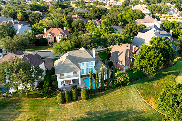 Aerial drone view of luxury mansions with swimming pools surrounded by green grass and trees in the summertime.